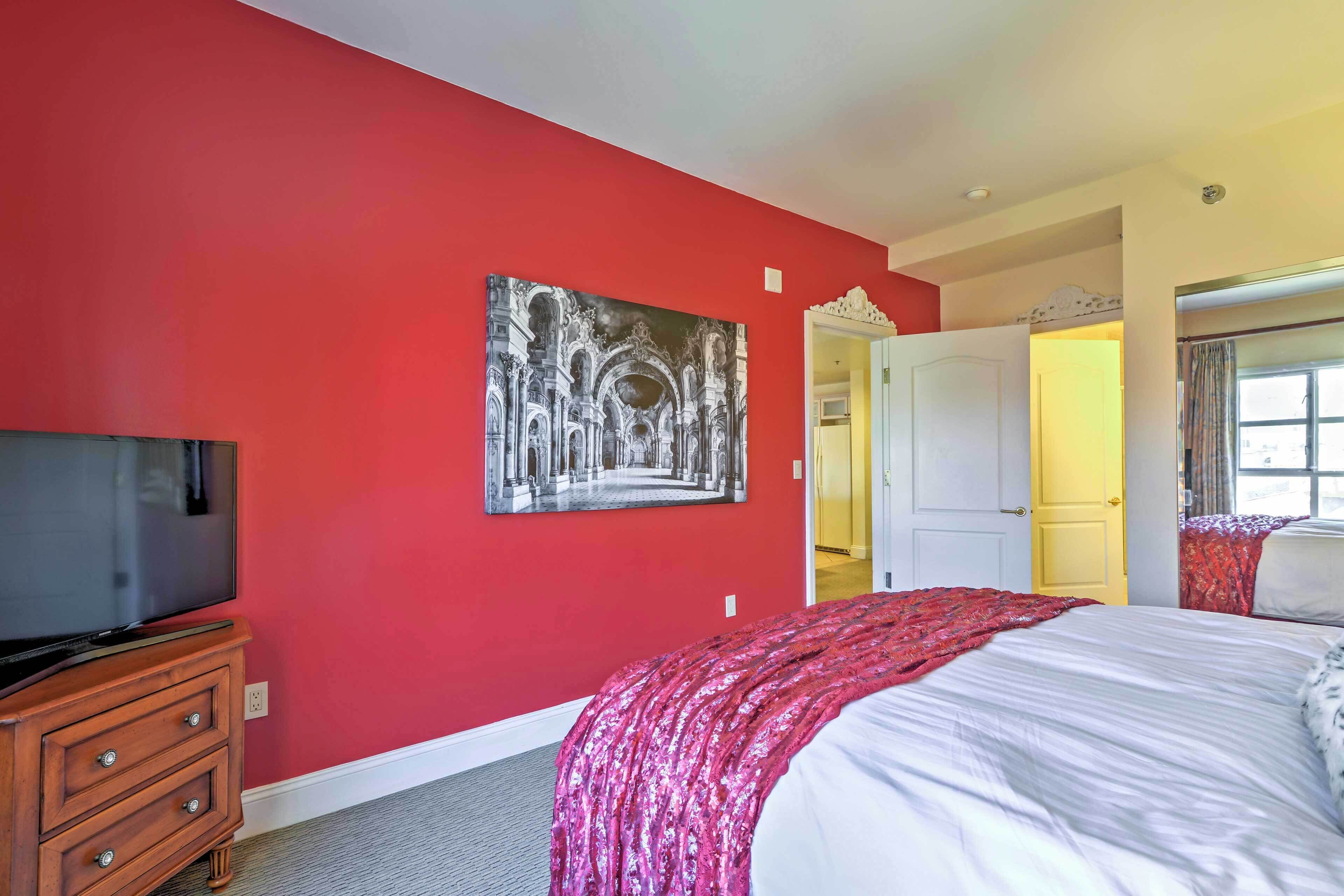 Un dormitorio con una pared roja, una cama con colcha blanca y rosa, un televisor de pantalla plana sobre una cómoda de madera y un cuadro de un edificio en la pared.