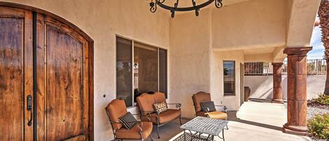 A covered patio with a wooden door, two cushioned chairs, a metal table, and a chandelier.