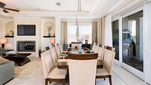 A dining area with a long table, cushioned chairs, a fireplace, and a view of the outdoor kitchen. - Lively Home with Water Views at Reunion (Kissimmee)