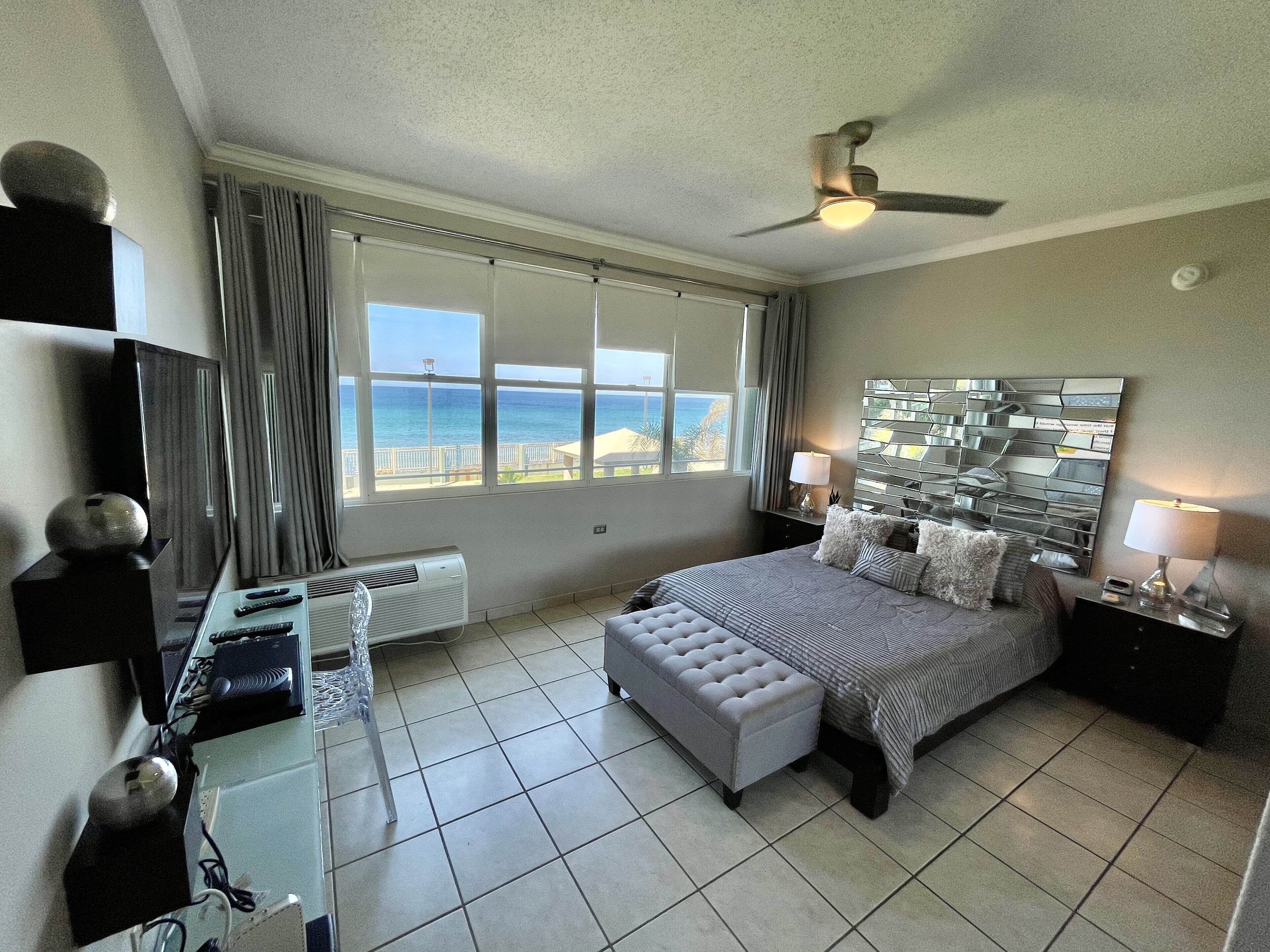 A hotel room with a bed, a desk with a computer, a TV, and a view of the ocean.