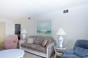 A living room with a patterned sofa, a pink armchair, a blue armchair, a white table, and two lamps. - Cozy, comfortable 2 bedroom oceanfront condo with coastal decor, free WiFi, and gorgeous view of the ocean and dunes just steps to the beach! (Ocean City)