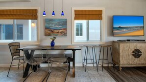 A dining area with a table, chairs, and a vase of flowers. There are two windows with blinds, a painting on the wall, and a television on the right. - Beautiful newly remodeled upper unit located within the first block to beach! (Newport Beach)