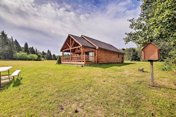 A rustic log cabin with a porch, surrounded by a grassy field and trees.