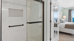 Une chambre d’hôtel avec une douche en verre, un lit avec du linge de lit blanc et une vue sur la mer depuis la porte ouverte.