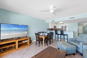 A living room with a TV, dining table, and chairs, a kitchen area with cabinets and appliances, and a blue couch.
