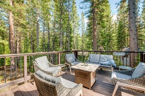 Relax on the deck surrounded by tall pine trees, with wicker chairs and a small table.