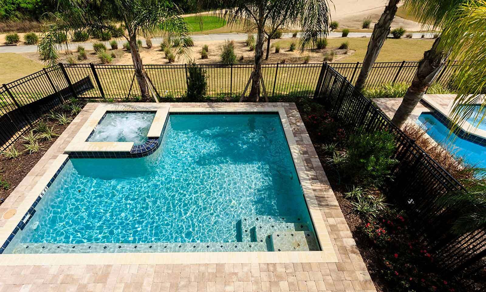 Une piscine rectangulaire avec un jacuzzi, entourée d’une terrasse en briques et d’une clôture en métal noir.
