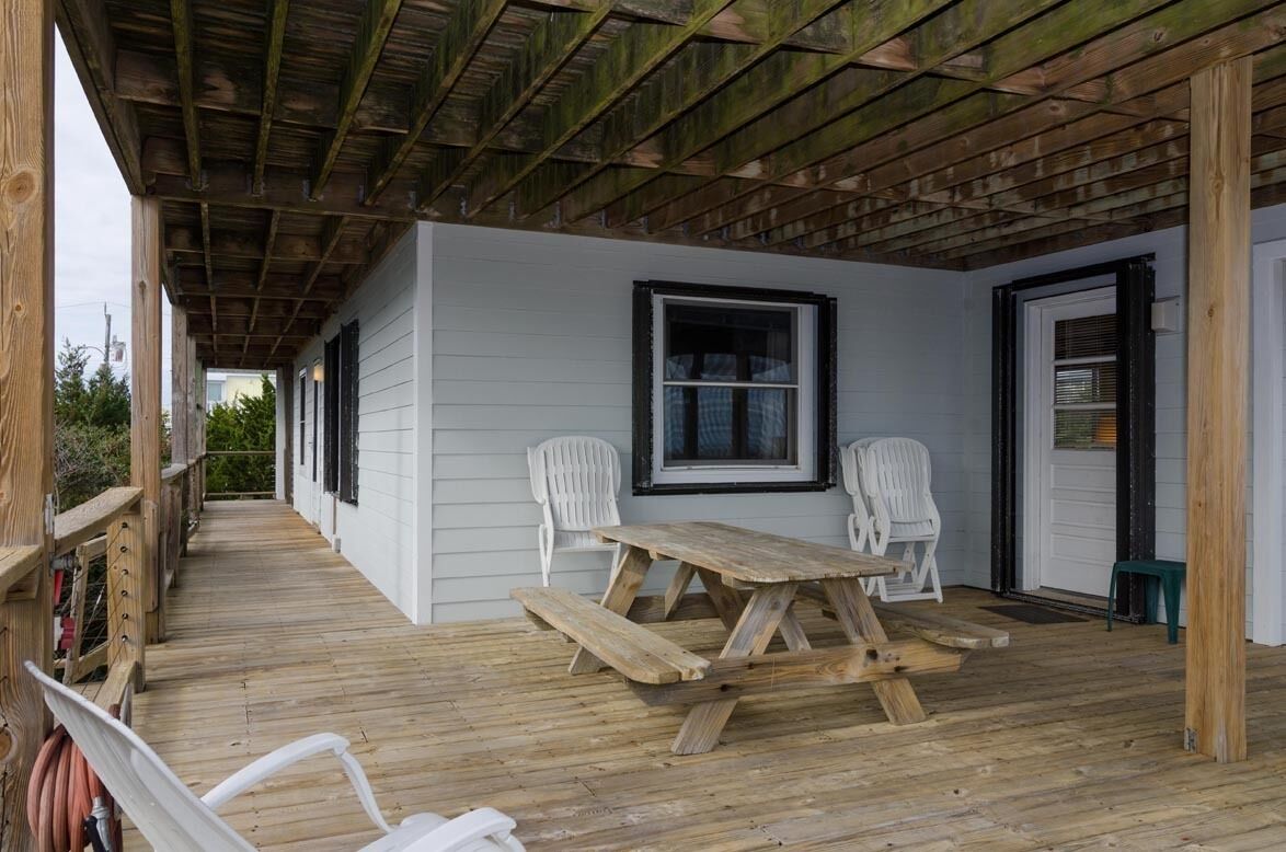 A covered deck with a picnic table and white chairs, wooden beams overhead, and a white building in the background.