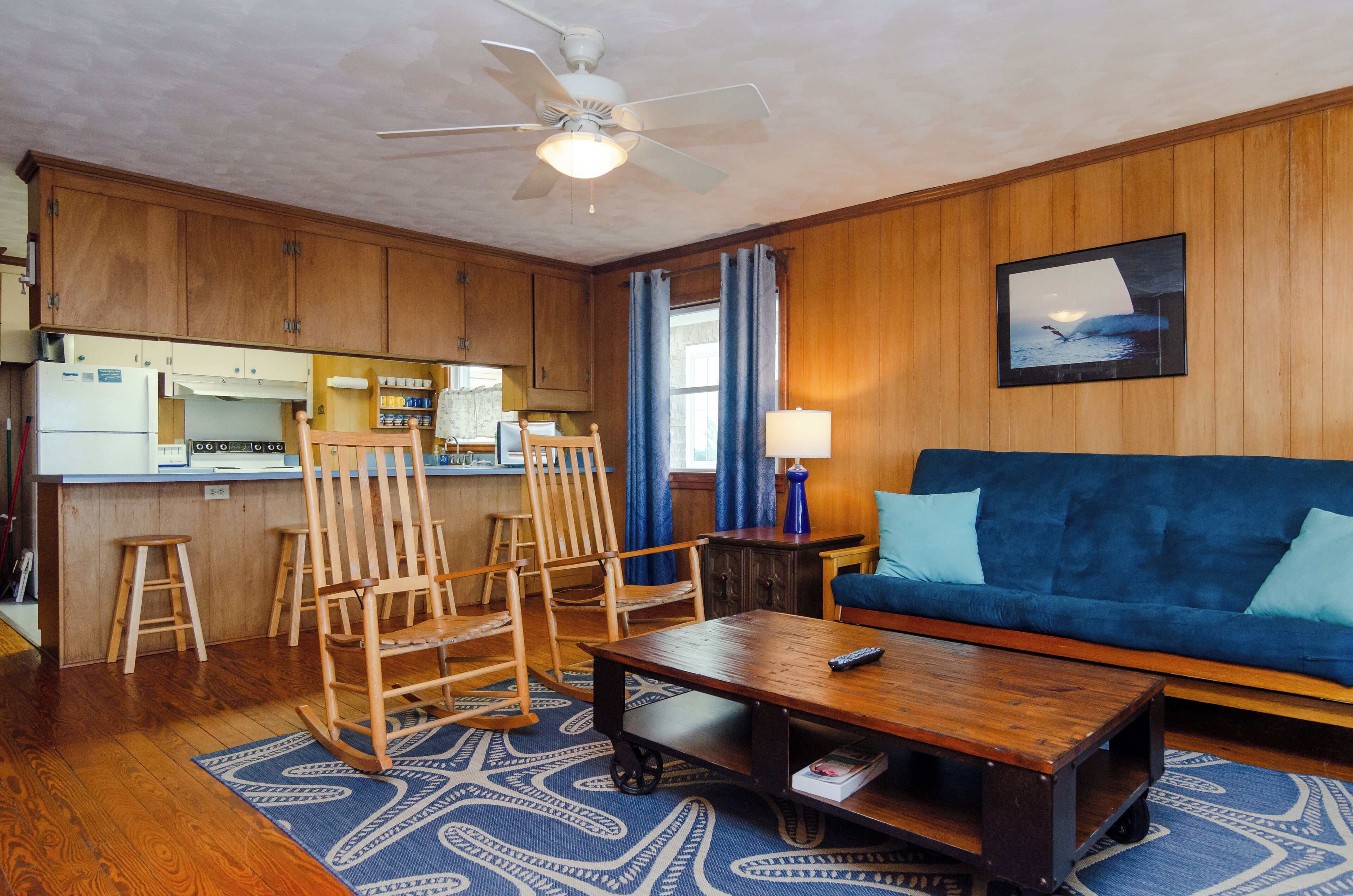 A living room with a blue sofa, wooden rocking chairs, a coffee table, and a kitchen area with wooden cabinets and a refrigerator.
