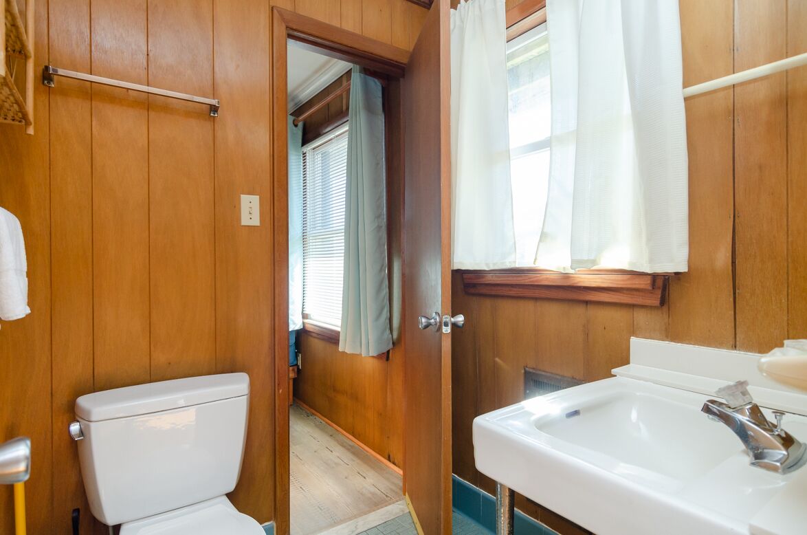 Bany amb panells de fusta, un vàter blanc, un lavabo i una finestra amb cortines.