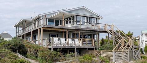 A two-story beach house with a balcony, a staircase, and a view of the beach.