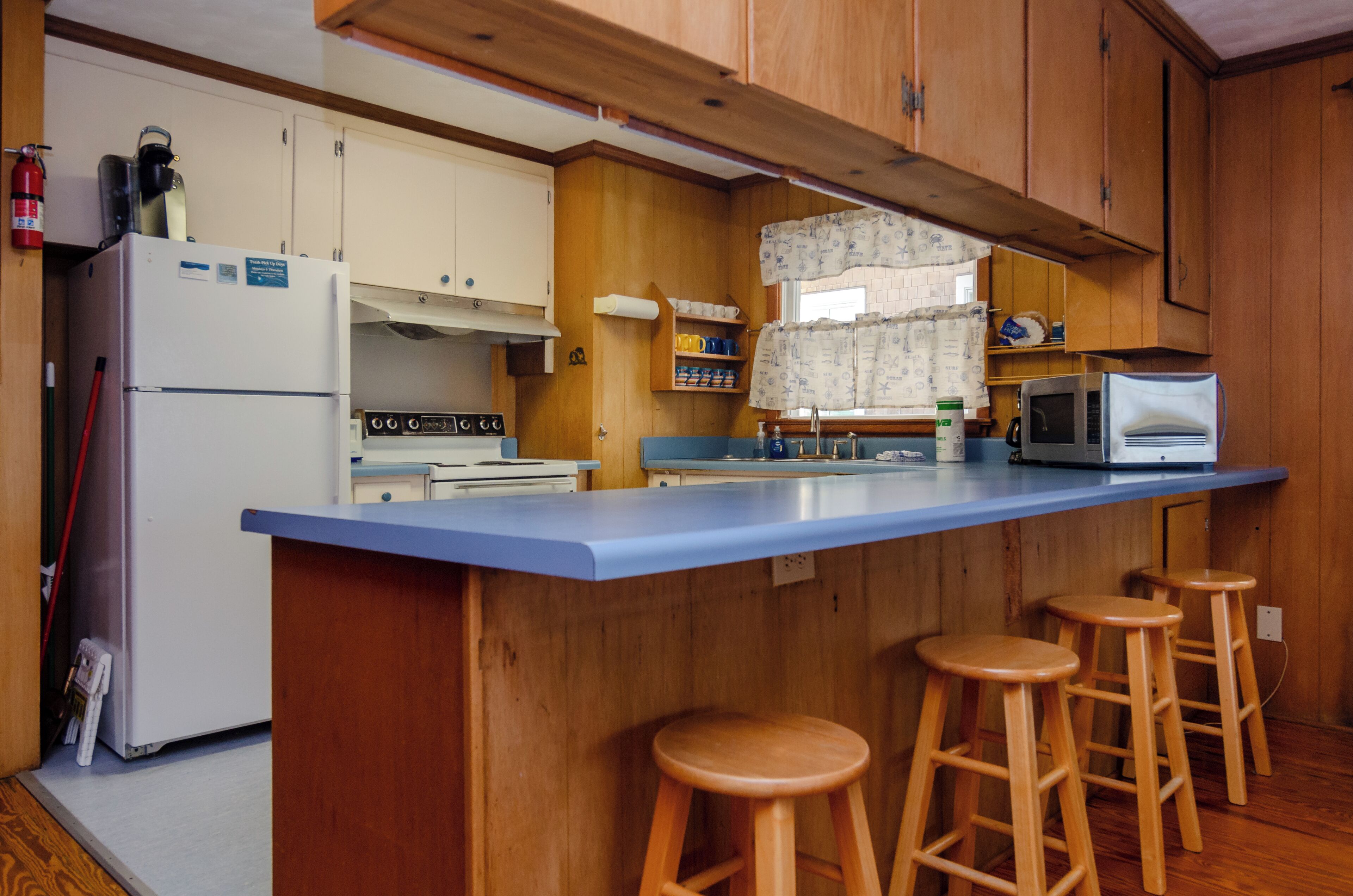 A compact kitchen with wooden cabinets, a white refrigerator, a stove, a microwave, and a blue countertop. There are three wooden stools.