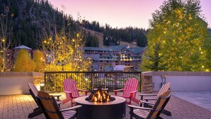 A circular outdoor seating area with a fire pit, surrounded by Adirondack chairs, overlooking a mountainous landscape with a lodge in the background.