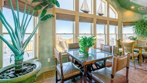 Un coin repas avec une table et des chaises en verre, de grandes plantes en pot et une vue sur des voiliers à travers de grandes fenêtres.