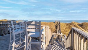 Une terrasse en bois avec des chaises blanches donnant sur une plage et des dunes.