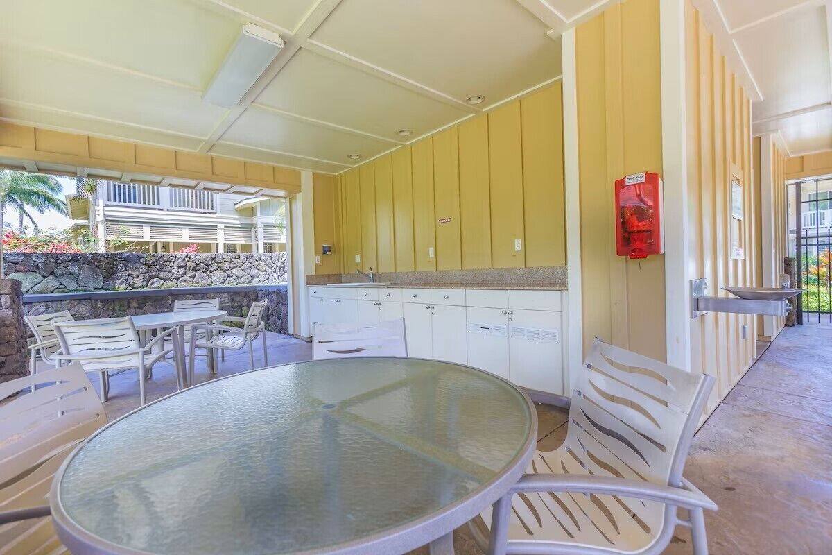 Een buitenpatio met een ronde glazen tafel en witte stoelen, een stenen muur en een gele muur met een rode brandblusser.