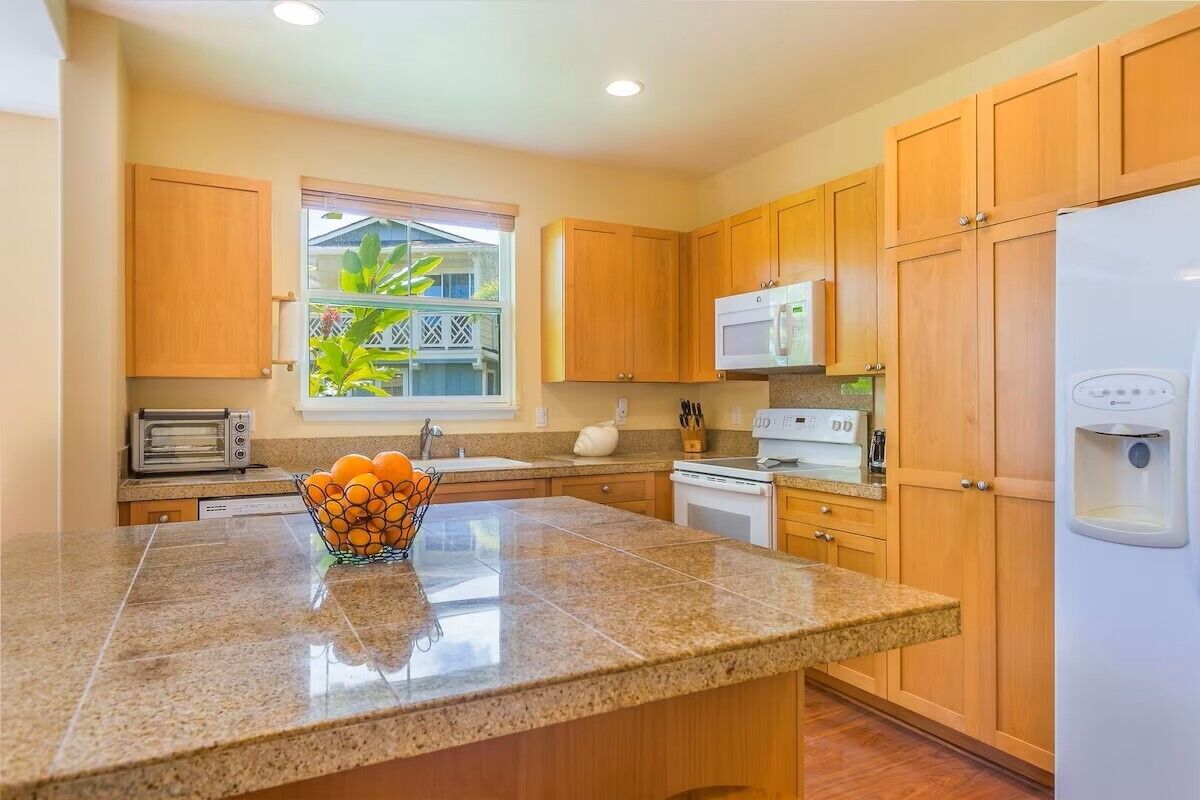 A kitchen with granite countertops, wooden cabinets, a microwave, a refrigerator, a toaster, and a window with a view of greenery.