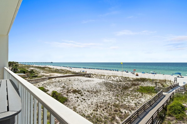 A beach view from a balcony with a wooden deck, sandy beach, and a pier in the distance.