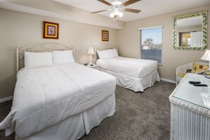 Quarto com duas camas, um ventilador de teto, janela com persianas, espelho e uma mesa pequena com abajur.