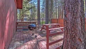 Una terraza de madera con mesas de picnic y una parrilla, rodeada de altos árboles.