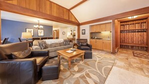 A spacious living room with a large sofa, a coffee table, and a fireplace. There is a kitchen area with wooden cabinets and a dining table.