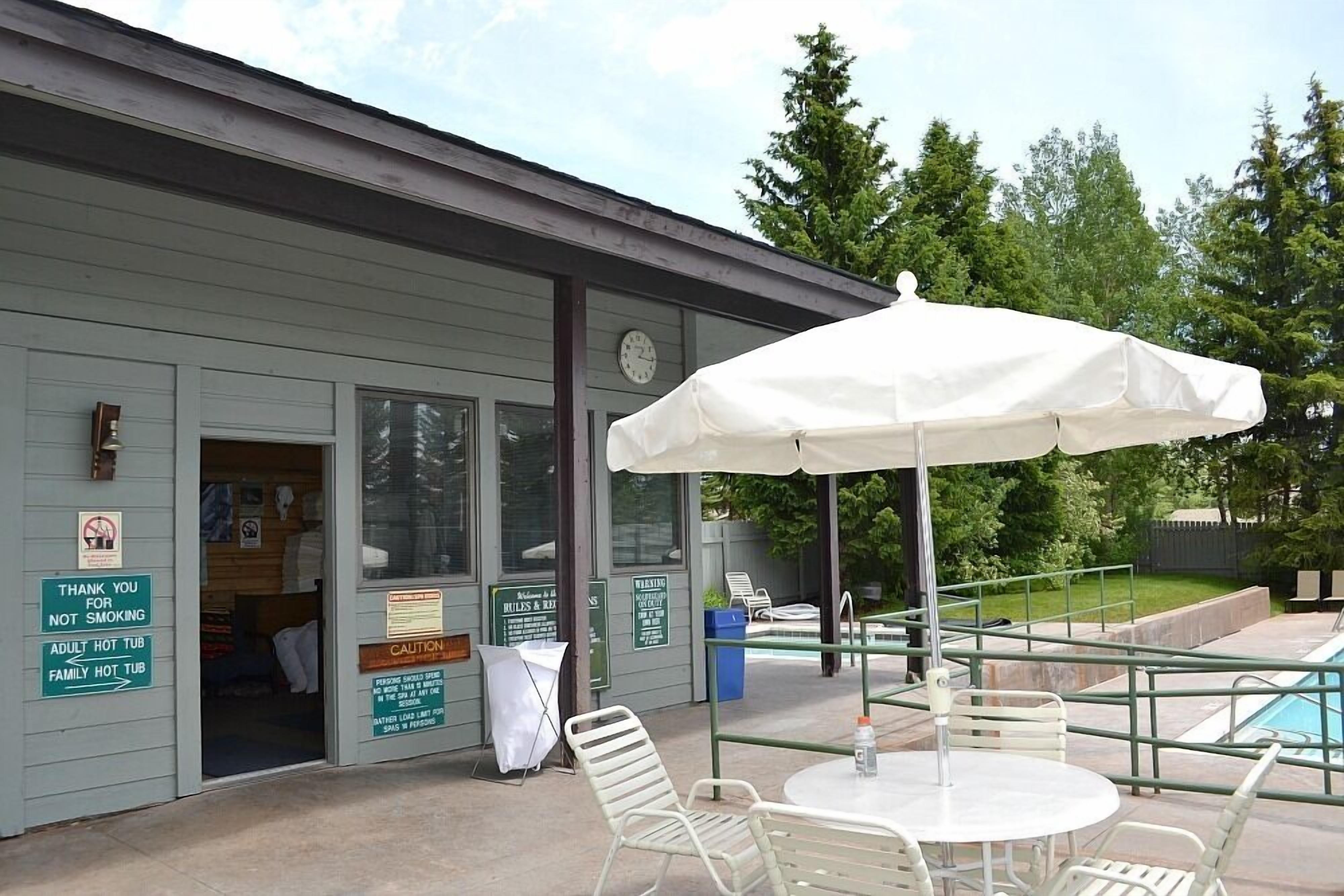 Un espace extérieur aménagé avec une terrasse, une piscine, un coin salon couvert avec un parasol blanc, et un bâtiment sur lequel sont affichés des panneaux indiquant les zones où il est interdit de fumer et la présence d’un jacuzzi.