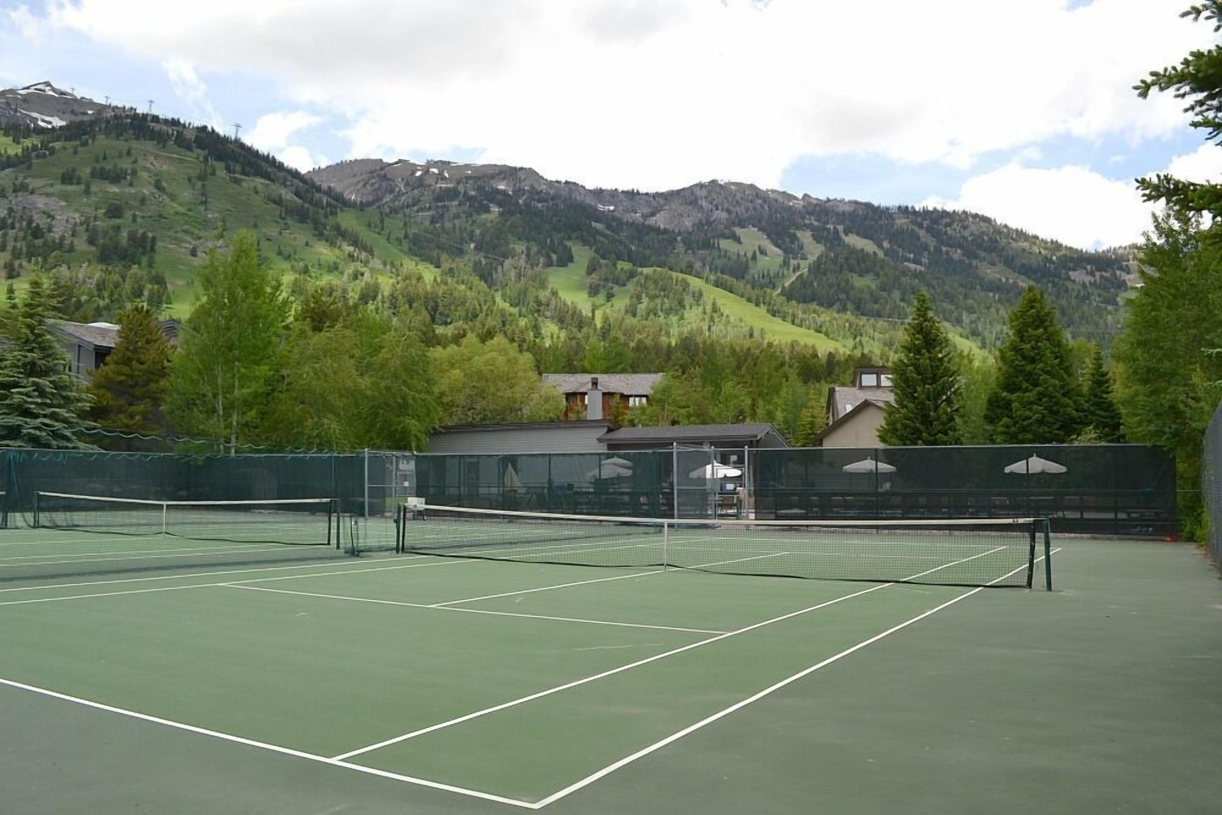 Un court de tennis avec un filet, entouré d’une clôture, et des bâtiments en arrière-plan.
