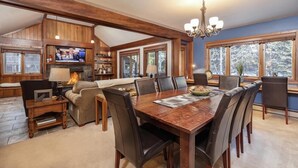 A spacious living area with a wooden dining table, comfortable seating, a fireplace, and large windows offering a view of the outdoors.