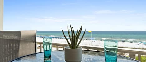A glass table with a potted plant and two glasses of blue liquid, overlooking a beach with umbrellas and people.