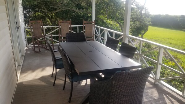 A covered outdoor dining area with a wooden table and wicker chairs, surrounded by a white railing with a view of a green field.