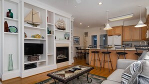 A modern living room with a fireplace, a television, and a coffee table. There is a kitchen area with bar stools and a refrigerator. - Charming 3 BR townhouse with sound views! (Wrightsville Beach)