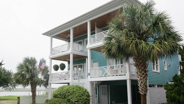 A two-story building with a balcony on the upper floor, a garage, and a palm tree in the foreground. - Charming 3 BR townhouse with sound views! (Wrightsville Beach)