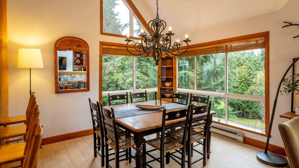 A dining area with a large table, chairs, a chandelier, and a view of the forest through large windows. - Spacious 3.5BR Retreat | Golf Course Views | Walk to Lakes & Trails | Managed By Aloha Whistler (Whistler)