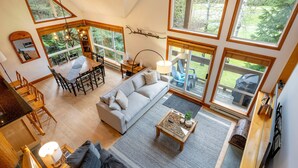 A spacious living room with a large sofa, dining table, and chairs, large windows offering a view of the outdoors. - Spacious 3.5BR Retreat | Golf Course Views | Walk to Lakes & Trails | Managed By Aloha Whistler (Whistler)