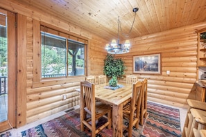 Un espace repas aménagé dans une cabane en bois, avec une grande table, des chaises, un lustre, une plante en pot et un tableau encadré au mur.