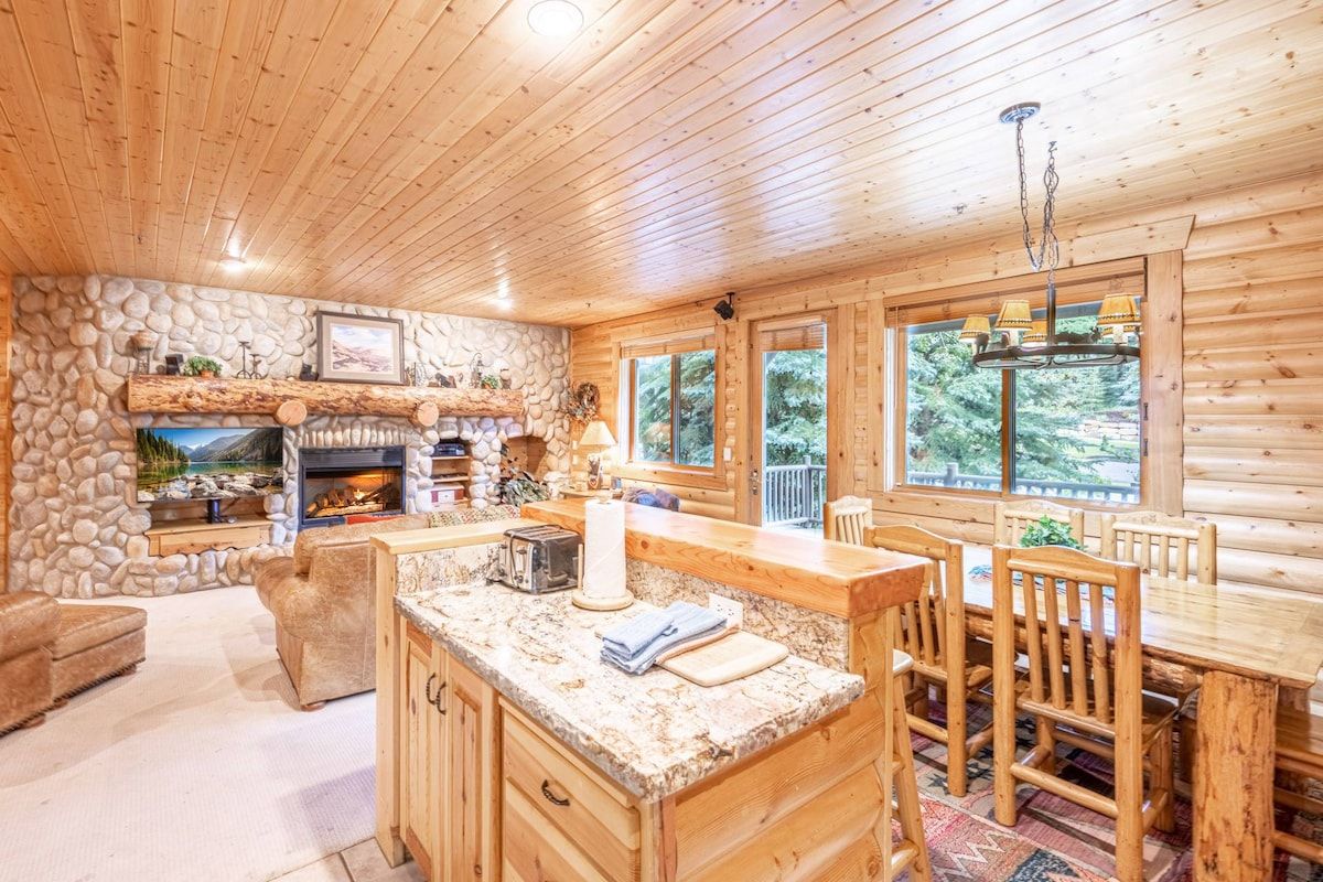 A log cabin interior with a stone fireplace, wooden dining table, and a large window.