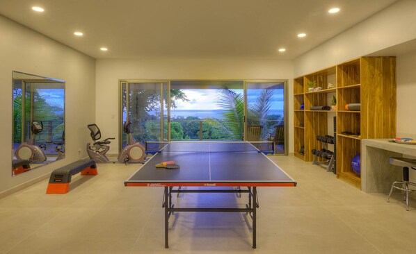 Une table de ping-pong dans une pièce dotée de grandes fenêtres, d’appareils de musculation et d’une étagère en bois.