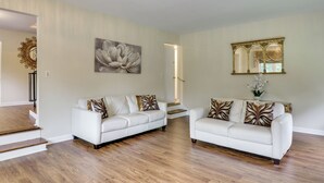 A spacious living area with two white sofas, a large floral painting, and a mirror with an ornate frame.