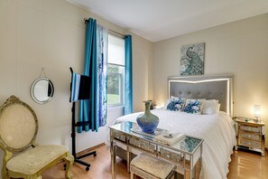 Une chambre à coucher comprenant un lit, un téléviseur, un miroir décoratif, une chaise, une table avec un vase et une table de chevet avec une lampe.