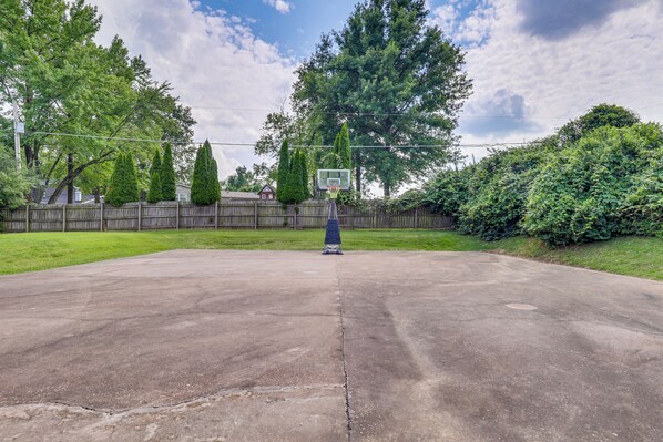 An outdoor basketball court with a hoop, surrounded by a wooden fence, trees, and a grassy area.
