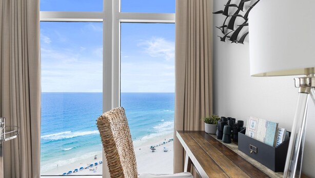 Una habitación con un ventanal grande con vista a la playa, una silla de mimbre, un escritorio de madera con una lámpara y un mueble negro con objetos encima.