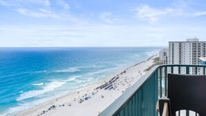 Vista de la playa desde un balcón con barandilla, el océano, sombrillas y un edificio de hotel al fondo.