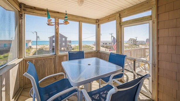 A covered patio with a table and chairs, large windows offering a view of the beach and ocean, and a wooden ceiling. - Bali Hai - Kitty Hawk MP 3, 50 Yards to Beach, Breathtaking Views, Open Decks (Kitty Hawk)