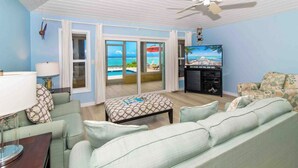 A living room with a view of the ocean, a ceiling fan, and a television. - Thatch Hill: Beach House w/ Private Pool & Backyard Snorkeling, Walk to the Rum Point Club! (Grand Cayman)