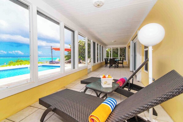 A sunlit balcony with wicker lounge chairs, a table with a fruit basket, and a view of a swimming pool and ocean.