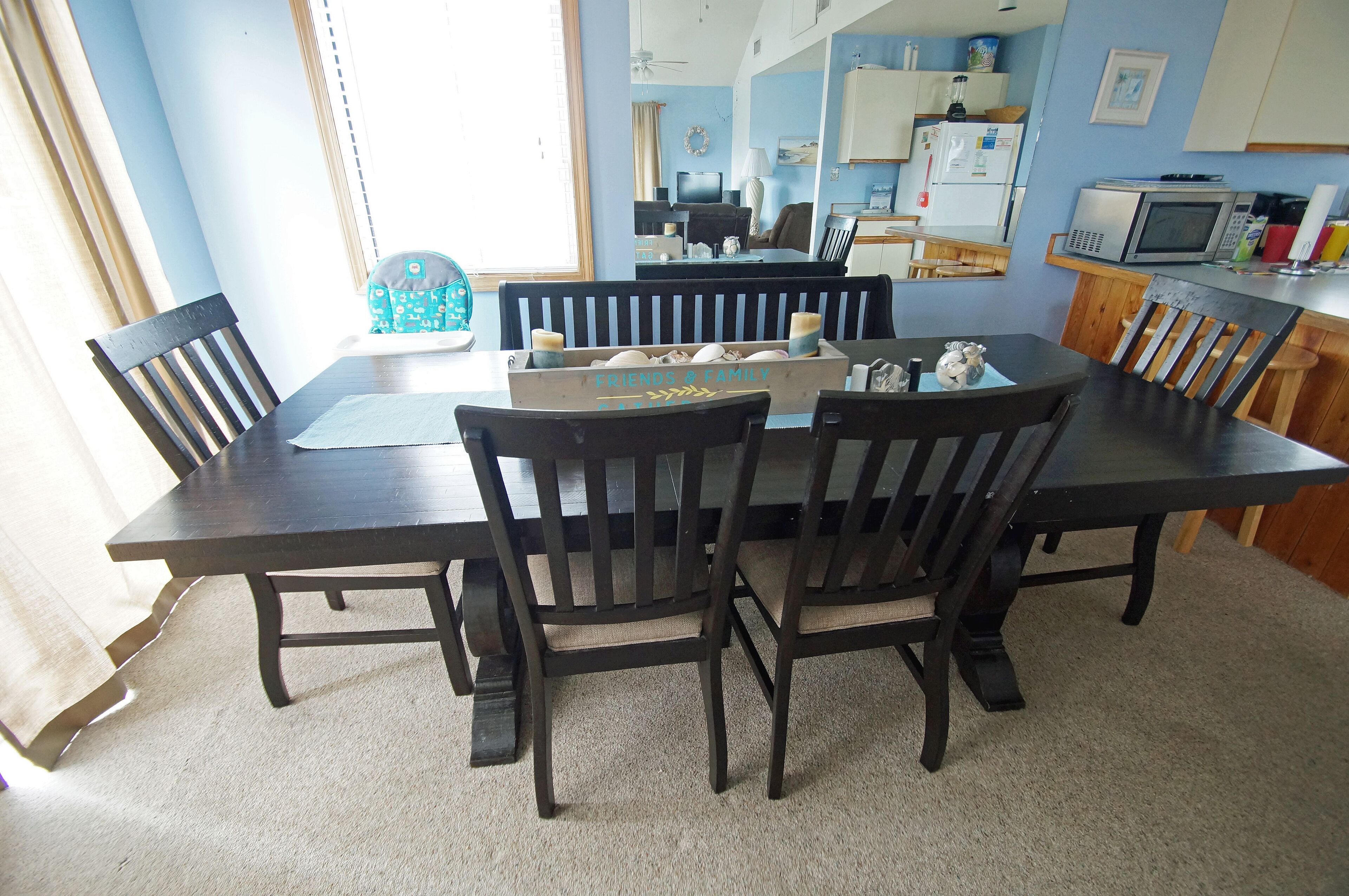 A dining area with a dark wooden table and chairs, a box labeled "FRIENDS A CAMPY" on the table, and a kitchen in the background.