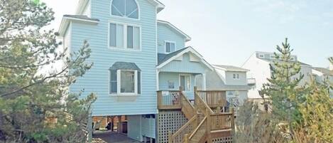 A two-story beach house with a wooden deck and a staircase leading to the entrance.