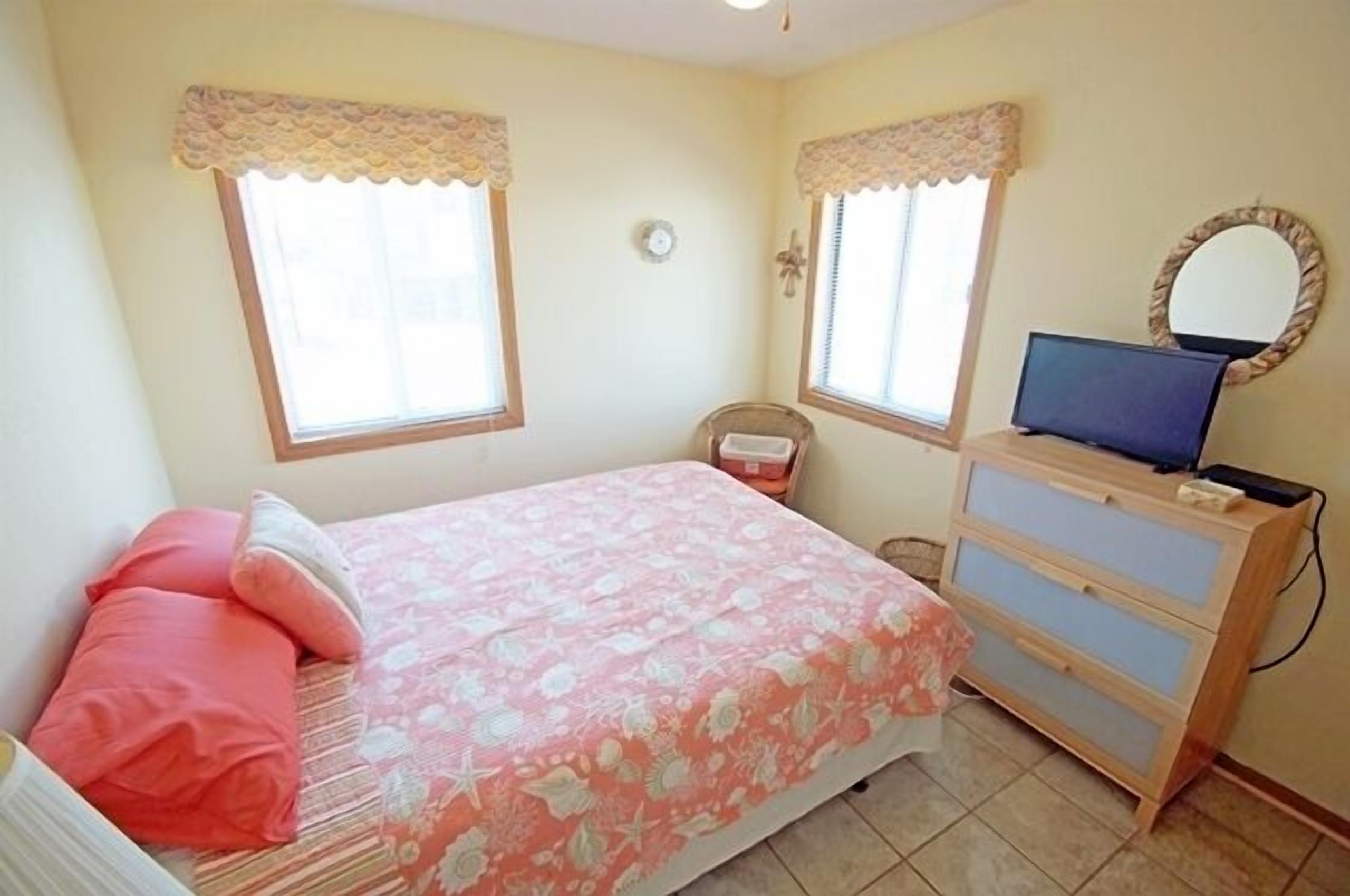 Een slaapkamer met een bed, een dressoir, een televisie en twee ramen met gordijnen.
