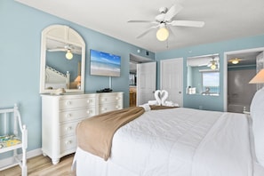 A bedroom with a bed, a dresser, a mirror, a ceiling fan, and a view of the beach.