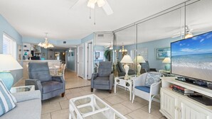 A spacious living room with a large flat-screen TV, comfortable seating, and a view of the ocean through a large window.